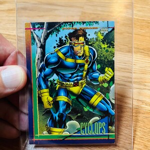 May include: A Cyclops trading card from the Marvel Super Heroes series, featuring the character in a dynamic pose. The card is in a protective sleeve. The card has a blue, yellow, and green color scheme.