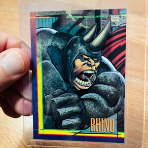 May include: A Rhino trading card featuring a detailed illustration of the character with a menacing expression. The card has a blue, yellow, and red border, and the name "RHINO" is prominently displayed at the bottom.