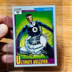 May include: A Marvel 1991 trading card featuring a character in a black and blue suit holding a weapon. The card has the text "Ultimate Nullifier" and is in a protective sleeve.