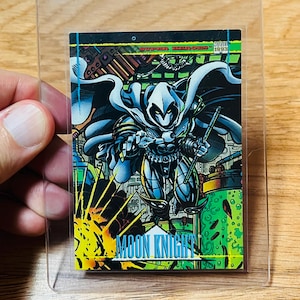 May include: A Moon Knight trading card featuring a comic book illustration of the character in a white hooded costume, holding weapons. The card has a colorful background with the words "MOON KNIGHT" at the bottom. The card is in a clear protective sleeve.