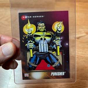 May include: A Punisher trading card from Marvel's 1992 Super Heroes series. The card features a detailed illustration of the Punisher holding two weapons with yellow flames. The card is encased in a clear plastic Card Saver I holder.
