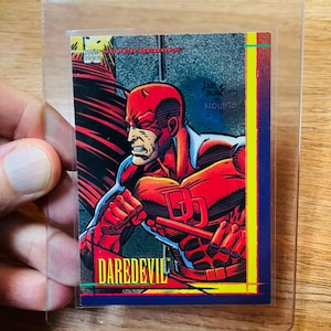 May include: A Daredevil trading card in a protective sleeve. The card features a comic book illustration of Daredevil in a red suit with horns. The card has the word "DAREDEVIL" in yellow text.