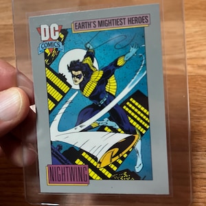 May include: A DC Comics Nightwing trading card in a protective sleeve. The card features a dynamic illustration of Nightwing in a blue and black costume, set against a cityscape background. Text includes "DC Comics", "Earth's Mightiest Heroes", and "Nightwing".