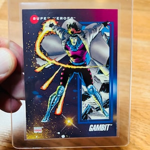 May include: A 1992 Marvel "Gambit" trading card in a protective sleeve. The card features a comic book illustration of Gambit, a superhero, with a dynamic pose and energy effects. The card has a blue, purple, and pink color scheme.