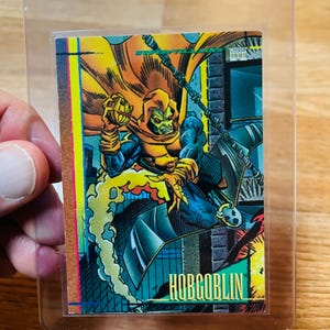 May include: A trading card featuring the Hobgoblin, a comic book character. The card shows the character in a dynamic pose, with a green and orange color scheme. The card is in a protective sleeve.