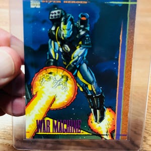 May include: A War Machine trading card from the Marvel Super Heroes 1992 series. The card features a full-body illustration of War Machine in a dynamic pose, with a blue and yellow color scheme, and a blast of energy.