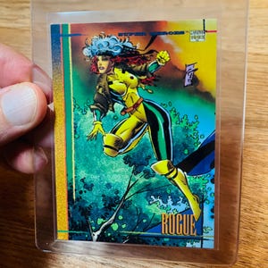 May include: A trading card featuring the character Rogue from the X-Men. The card has a colorful illustration of Rogue in a yellow and green costume, with the word "ROGUE" at the bottom. The card is encased in a clear plastic sleeve.