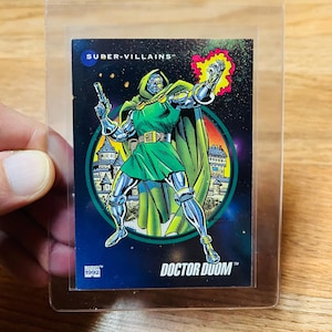 May include: A Doctor Doom trading card from the Super-Villains series. The card features a green-robed figure with a metal mask and armor, holding a gun and a burst of energy. The card is in a protective sleeve.