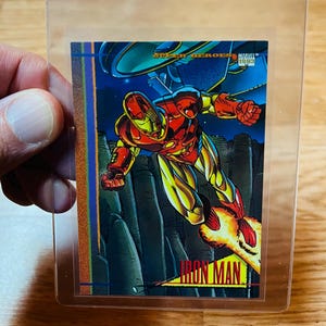 May include: A Marvel Super Heroes trading card featuring Iron Man in flight. The card has a vibrant illustration of Iron Man in his red and gold armor, with a blue and black background. The card is encased in a clear plastic sleeve.