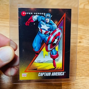 May include: A Captain America trading card from 1992, featuring the superhero in a blue, red, and white costume, holding his shield. The card is in a clear protective sleeve, with the words "SUPER HEROES" and "CAPTAIN AMERICA" visible.