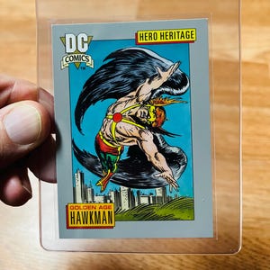 May include: A DC Comics trading card featuring Hawkman from the Golden Age. The card shows Hawkman in a dynamic pose with wings spread against a blue sky, with the text "HERO HERITAGE" and "HAWKMAN" visible.