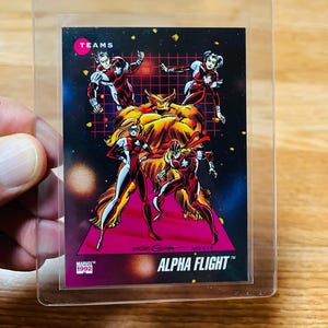 May include: A trading card featuring the Alpha Flight team from Marvel Comics. The card has a black background with a grid pattern and images of the characters in action poses. The card is in a protective sleeve. The text "ALPHA FLIGHT" is visible.