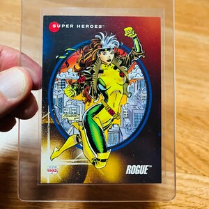 May include: A trading card featuring the Marvel Comics character Rogue, in a clear protective sleeve. The card has a colorful illustration of Rogue in a yellow and green costume, with the text "SUPER HEROES" and "ROGUE" visible. The card is from 1992.