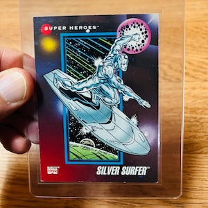 May include: A Silver Surfer trading card from 1992, featuring the character on a surfboard against a cosmic background. The card is in a protective sleeve, with the words "SUPER HEROES" and "SILVER SURFER" visible.