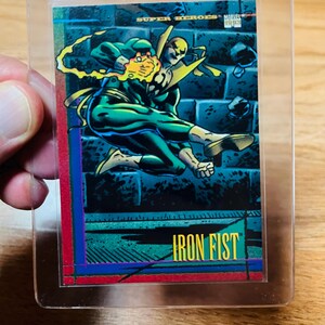 May include: A Marvel Super Heroes trading card featuring Iron Fist in action. The card has a vibrant design with a blue, green, and yellow color scheme. The card is encased in a clear plastic sleeve for protection.