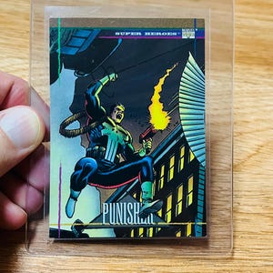 May include: A Marvel Super Heroes trading card featuring The Punisher in action. The card shows The Punisher in a black and blue costume, firing a gun with a yellow flame. The card is in a protective sleeve.