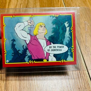 May include: A vintage trading card featuring He-Man from Masters of the Universe. The card shows He-Man with a sword, wearing a pink top and white sleeves, with the text "BY THE POWER OF GRAYSKULL..." in a speech bubble.