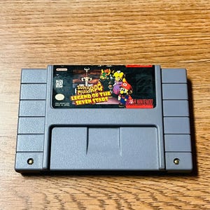 May include: A gray Super Nintendo cartridge for the game "Super Mario RPG: Legend of the Seven Stars." The label features colorful artwork of Mario, Bowser, and other characters. The cartridge is rectangular with a series of ridges and a small screw on each side.