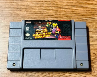 May include: A gray Super Nintendo cartridge for the game "Super Mario RPG: Legend of the Seven Stars." The label features colorful artwork of Mario, Bowser, and other characters. The cartridge is rectangular with a series of ridges and a small screw on each side.
