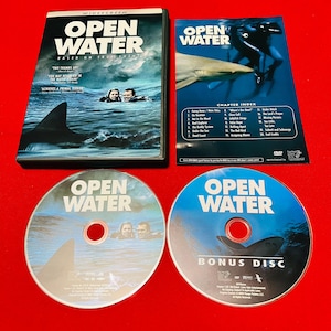 May include: A DVD set of the movie "Open Water" with a black case, two discs, and a chapter index. The cover features a shark fin and two people in the water. The discs have the movie title and images.