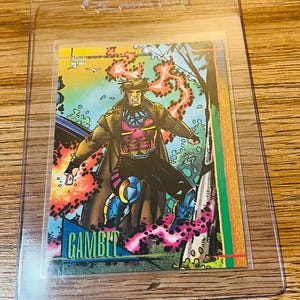 May include: A Gambit trading card featuring a comic book illustration of the character in a dynamic pose. The card has a colorful background with the character's name, "GAMBIT," at the bottom. The card is encased in a clear protective sleeve.