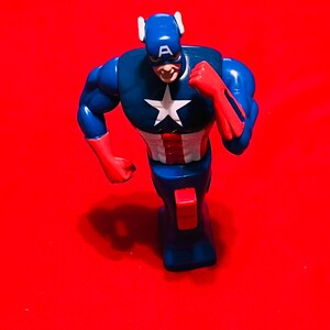 May include: A blue and red plastic action figure of Captain America running. The figure has a white star on the chest and a red, white, and blue design on the legs. The figure is on a red background.