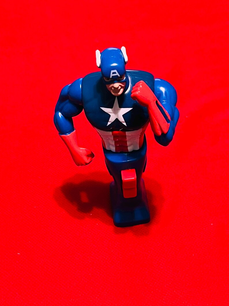 May include: A blue and red plastic action figure of Captain America running. The figure has a white star on the chest and a red, white, and blue design on the legs. The figure is on a red background.
