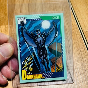 May include: A Marvel 1991 Darkhawk rookie trading card. The card features a superhero in a black and blue suit with outstretched wings, set against a cityscape and night sky. The card is in a protective sleeve.