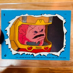 May include: A trading card featuring a cartoon character with a pink brain in a yellow and red helmet. The card has a blue background with a torn paper effect. The number "1" is in the upper left corner.