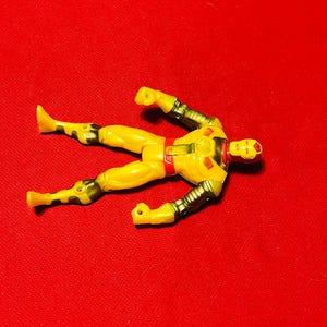 May include: Yellow action figure of a superhero with a red and gold chest piece and a red face mask. The figure is lying on its back with its arms and legs outstretched.