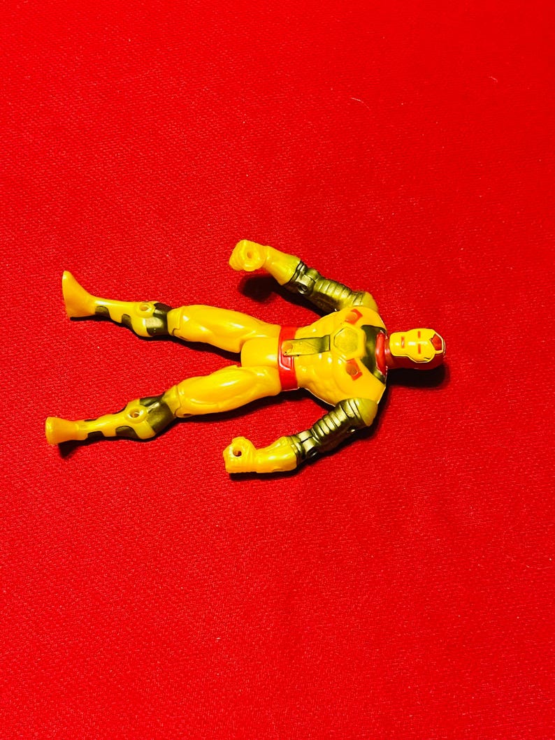 May include: Yellow action figure of a superhero with a red and gold chest piece and a red face mask. The figure is lying on its back with its arms and legs outstretched.