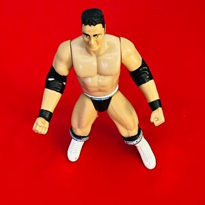 May include: A black and white wrestling action figure with black wrist bands and a white and black wrestling outfit. The figure has a muscular build and is posed in a fighting stance.