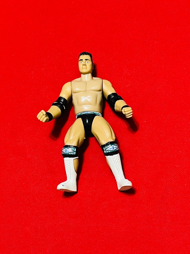 May include: A wrestling action figure with a black and white wrestling outfit. The figure has black wrist bands and white boots with black and blue accents. The figure has a muscular build and is posed in a fighting stance.