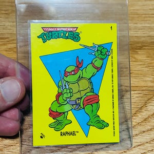 May include: A Teenage Mutant Ninja Turtles trading card featuring Raphael in a dynamic pose, holding two sais. The card has a yellow background with a blue triangle and the Turtles logo. The card is in a clear plastic sleeve.