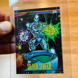 May include: A Silver Surfer trading card in a protective sleeve. The card features a metallic, muscular figure on a silver surfboard against a cosmic background. The text "SILVER SURFER" is prominently displayed.