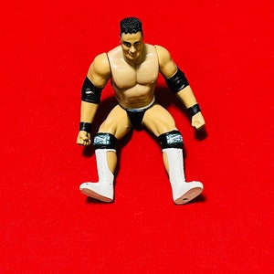 May include: A plastic action figure of a wrestler wearing black and white wrestling gear. The figure has a muscular build and is posed in a sitting position with legs spread apart. The figure is on a red background.
