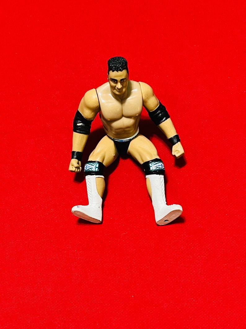 May include: A plastic action figure of a wrestler wearing black and white wrestling gear. The figure has a muscular build and is posed in a sitting position with legs spread apart. The figure is on a red background.