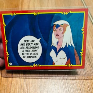 May include: A vintage trading card featuring a female character with white hair and a blue and white outfit. The card has a red border with a jagged design. Text in a speech bubble reads: "TRAP JAW AND BEAST MAN ARE ASSEMBLING A HUGE ARMY IN THE REGION OF STARDICA!"