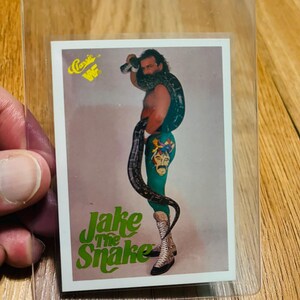 May include: A wrestling trading card featuring Jake "The Snake" Roberts. The card shows Roberts with a snake around his neck and leg. The card has the text "Jake the Snake" in green and the WWF logo.