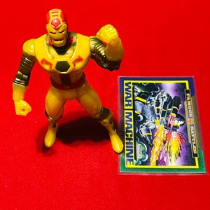 May include: Yellow and red plastic action figure of Iron Man's alter ego War Machine. The figure is posed with one arm raised and the other arm bent at the elbow. The figure is standing on a red surface. A trading card with the text "WAR MACHINE" is visible in the background.