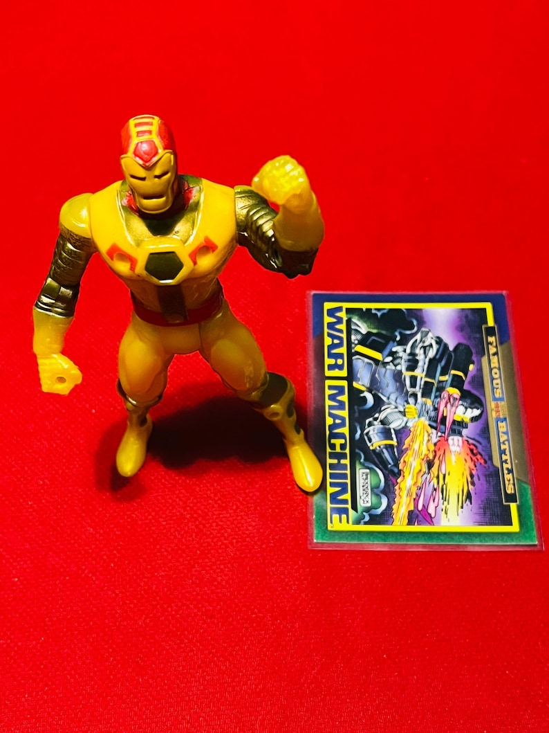 May include: Yellow and red plastic action figure of Iron Man's alter ego War Machine. The figure is posed with one arm raised and the other arm bent at the elbow. The figure is standing on a red surface. A trading card with the text "WAR MACHINE" is visible in the background.