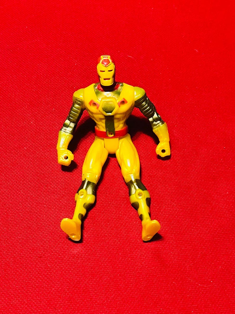 May include: Yellow and black action figure of a superhero with a red and yellow helmet. The figure has a black belt and is standing on a red background.