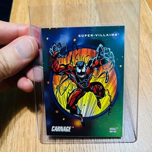 May include: A trading card featuring the Marvel Comics villain Carnage. The card has a colorful design with a red and black figure against a blue and green background. The text "CARNAGE" and "SUPER-VILLAINS" are visible.