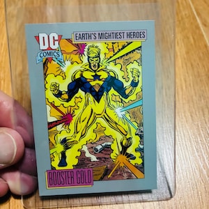 May include: A rectangular DC Comics trading card featuring the superhero Booster Gold in a yellow and blue costume, surrounded by yellow energy effects. The card includes the text Earth's Mightiest Heroes and Booster Gold, encased in a clear plastic sleeve.