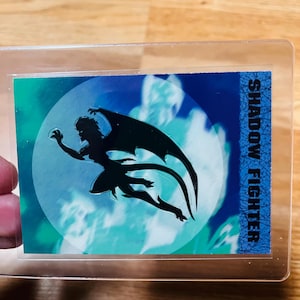 May include: A trading card featuring a silhouetted figure with large wings against a blue and green background. The card is encased in a clear plastic holder. The text "SHADOW FIGHTER" is printed vertically on the right side.