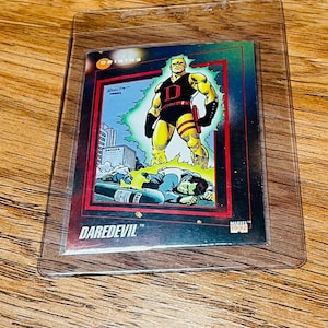 May include: A Daredevil trading card in a protective sleeve. The card features a comic book illustration of Daredevil in a black and yellow suit, standing over defeated figures. The card has a red border and the word "DAREDEVIL" at the bottom.