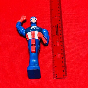 May include: A blue and red plastic action figure of Captain America standing approximately 5 inches tall. The figure is posed with one arm raised and the other arm bent at the elbow. The figure is wearing a blue and red costume with a white star on the chest.