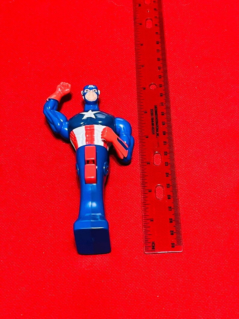 May include: A blue and red plastic action figure of Captain America standing approximately 5 inches tall. The figure is posed with one arm raised and the other arm bent at the elbow. The figure is wearing a blue and red costume with a white star on the chest.