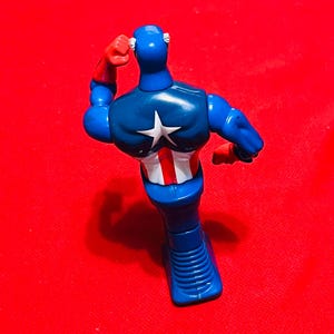 May include: A blue and red plastic action figure of Captain America standing with his arm raised. The figure is wearing a blue and red costume with a white star on the chest.
