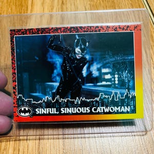 May include: A trading card featuring Catwoman in a black catsuit, with a cityscape background. The card has the Batman logo and the text "SINFUL, SINUOUS CATWOMAN". The card is in a protective sleeve.